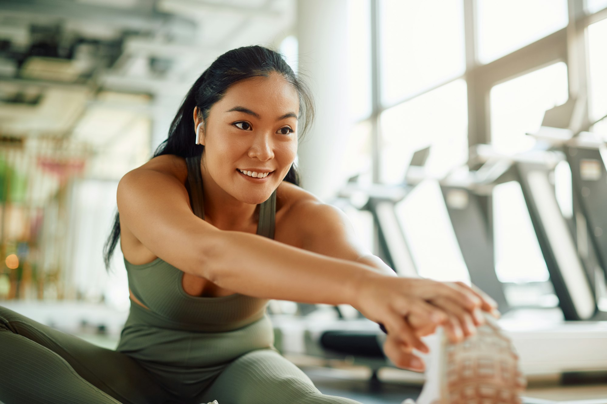 Happy Asian athlete stretching her leg while warming up for sports training in a gym